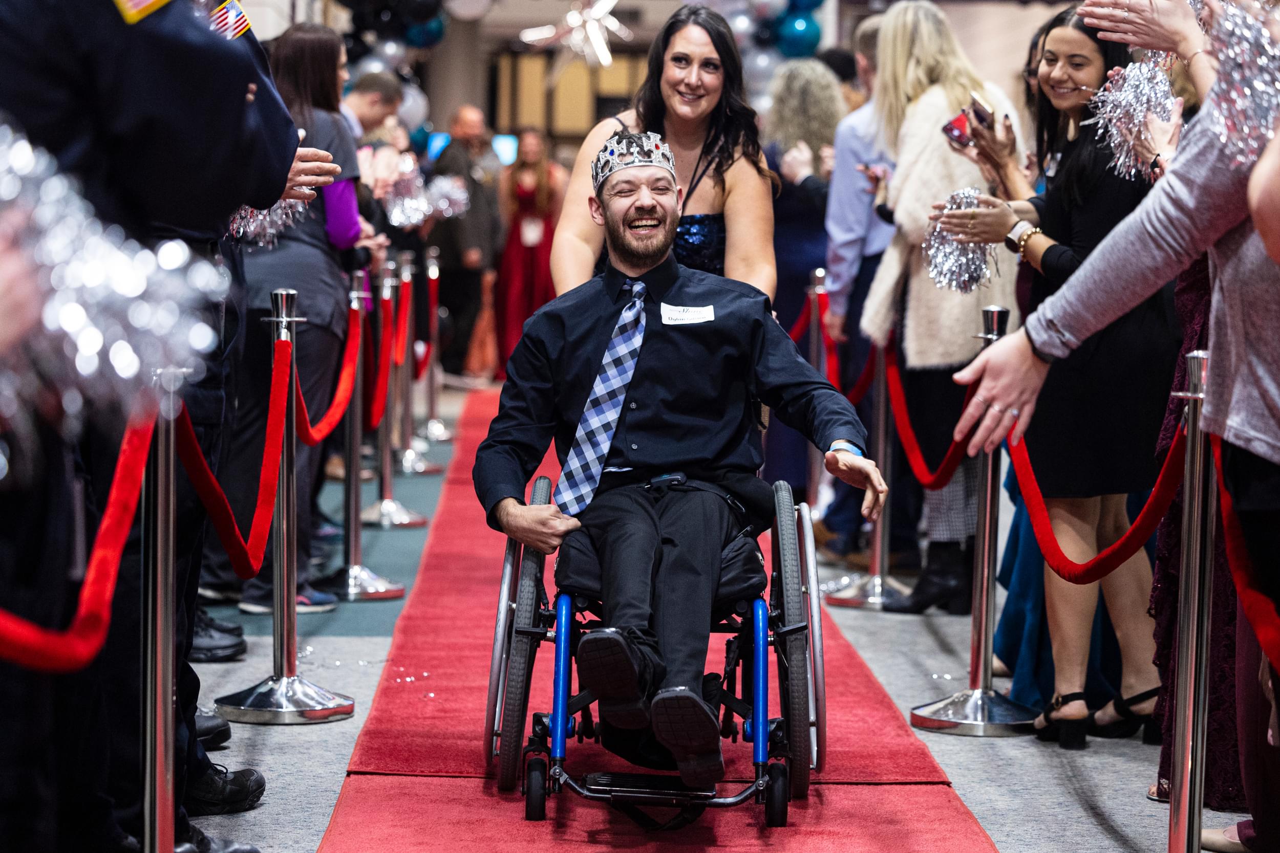 Man in wheelchair smiling as woman pushes him down the red carpet